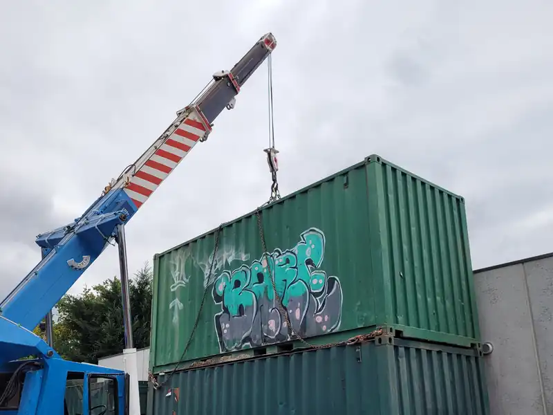 Two green shipping containers stacked, with a crane boom and hook beside them